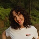 A cheerful woman with curly hair smiling outdoors in a green forest setting.